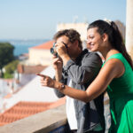 Portrait of loving couple taking photos of summer sea and city. Happy man and woman standing on city upland enjoying rest and man taking photos of faraway sea. Love, vacation and travelling concept