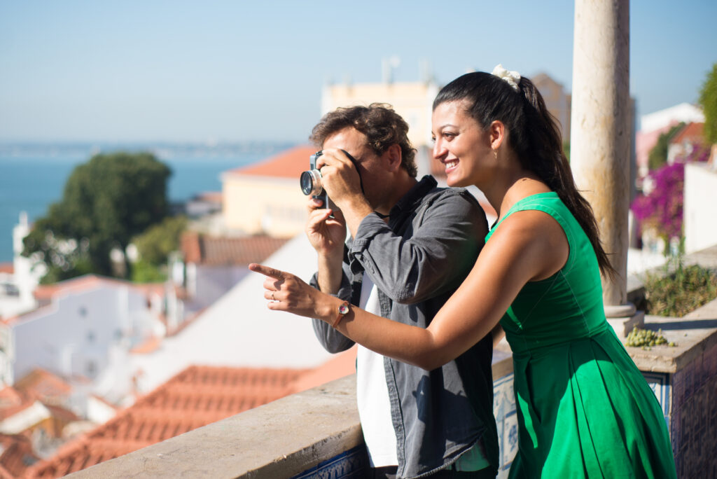 Portrait of loving couple taking photos of summer sea and city. Happy man and woman standing on city upland enjoying rest and man taking photos of faraway sea. Love, vacation and travelling concept